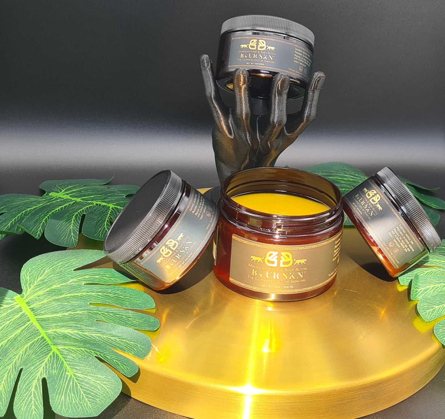 Vegan Mahogany Body & Hair Butter jars, handcrafted for skin and scalp, displayed on a gold pedestal with green leaves.